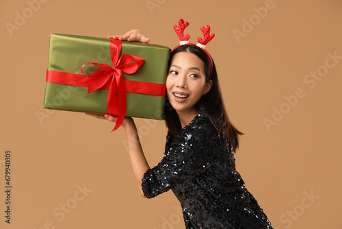 Young Asian woman in reindeer horns with Christmas gift box on brown background