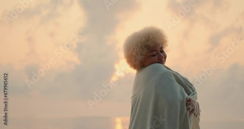 A young African American woman with curly blond hair is wrapped in a blanket and baskin near the ocean at dawn. Cozily wrapped in a warm blanket on the beach. Meet the first rays of the sun. photo