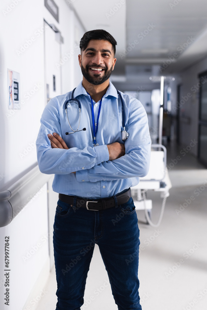 Portrait of happy biracial male doctor with stethoscope standing in hospital corridor