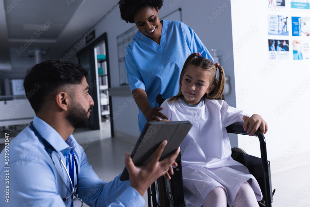 Diverse male and female doctors talking with girl in wheelchair using tablet in hospital corridor Diverse male and female doctors talking with girl in wheelchair using tablet in hospital corridor