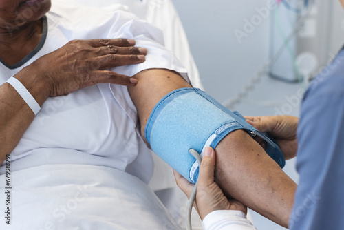 Diverse female doctor testing blood pressure of senior female patient in hospital room