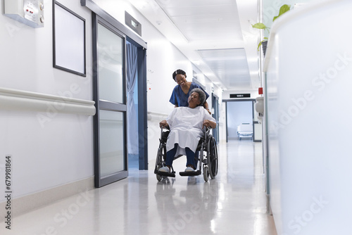 African american female doctor walking with senior female patient in wheelchair in hospital corridor