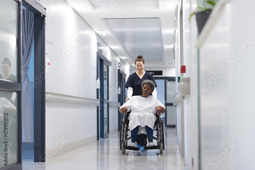 Diverse female doctor walking with senior female patient in wheelchair in hospital corridor