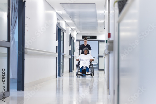 Diverse female doctor walking with senior female patient in wheelchair in hospital corridor