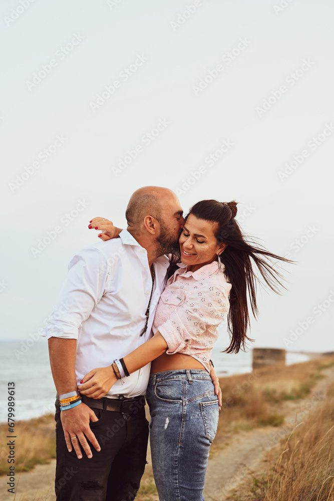 Happy smiling  Couple drinking vine at Sand Dunes near the Beach.  Young happy Bearded muscular  man  in White shirt kissing and hugging beautiful woman at sunset on a beach
