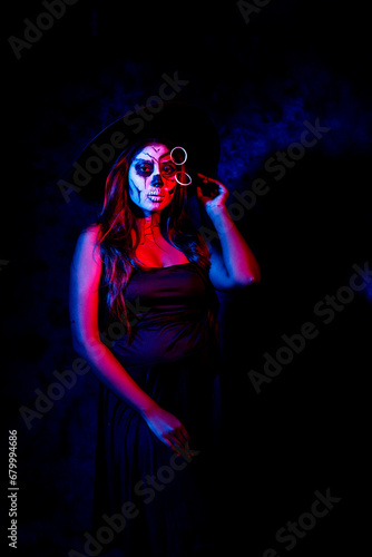 A women with Halloween Makeup in blue red light giving out a horror look wearing black gown in black background © Kaushik