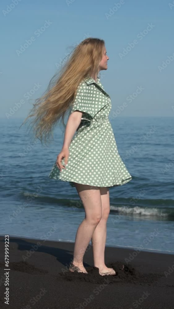 Authentic female with long hair in summer green polka dot dress playful spinning around herself on beach of Pacific Ocean. Sensual blonde woman with long legs barefoot. Handheld. Vertical slow motion