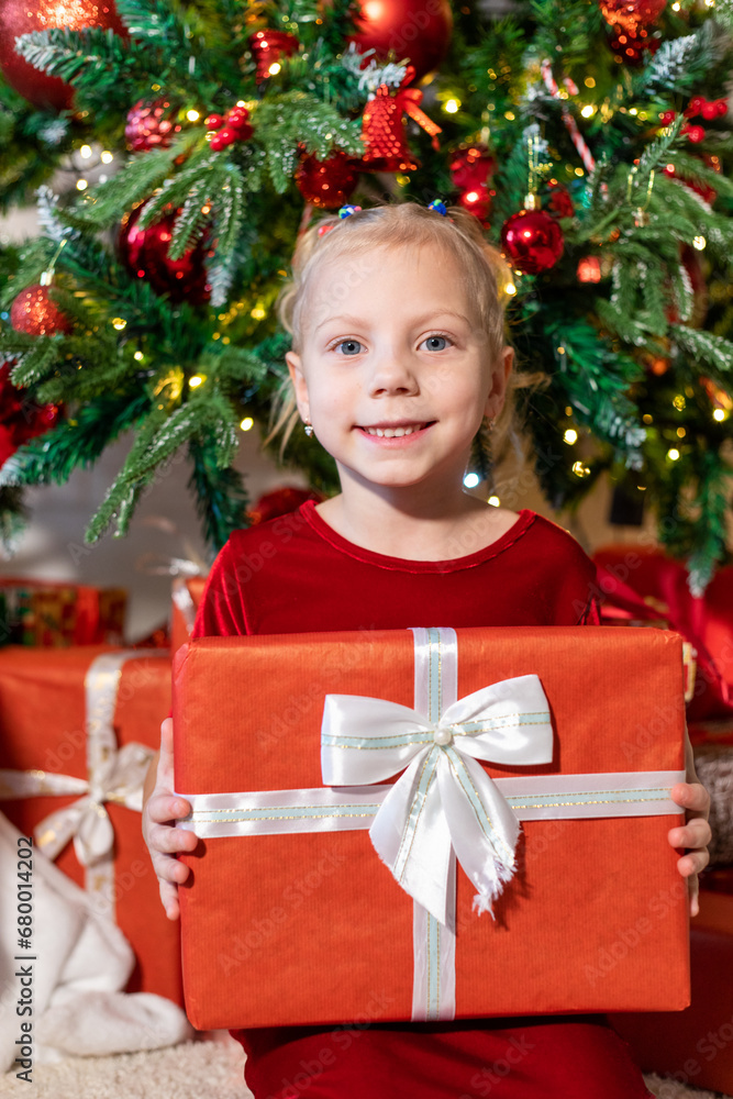 Cute girl with a gift in her hands sits near the Christmas tree.