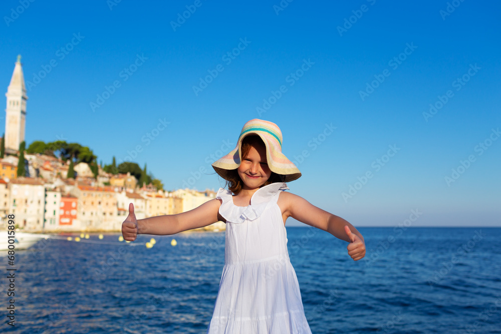 Funny little girl posing on embankment sunny summer day. Rovinj town in background. Travel and adventure concept.