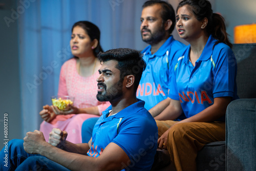 Tensed Senior Middle aged parents with siblings watching live Cricket sports match with Indian Tshirts at home on sofa - concept of entertainment  Nail biting and intense expression.