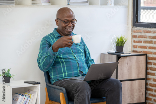 Senior black man relaxing from work. Sip hot drink and watch entertainment on laptop screen in office's relaxing corner.