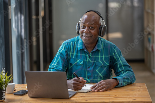 Senior black man wearing headphones enjoying listening to music or something interesting to relax while working. Study and take notes of interesting and useful things.