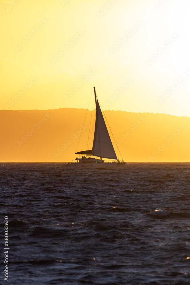 Yacht sailing in the Aegean sea in front of mountainous island filled by vivid orange sunset light