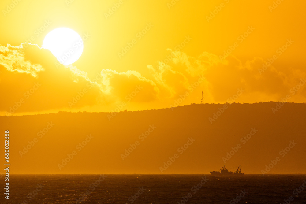 A beautiful sunset with a silhouette of the sun and a small ship in a background of a sea and the sun