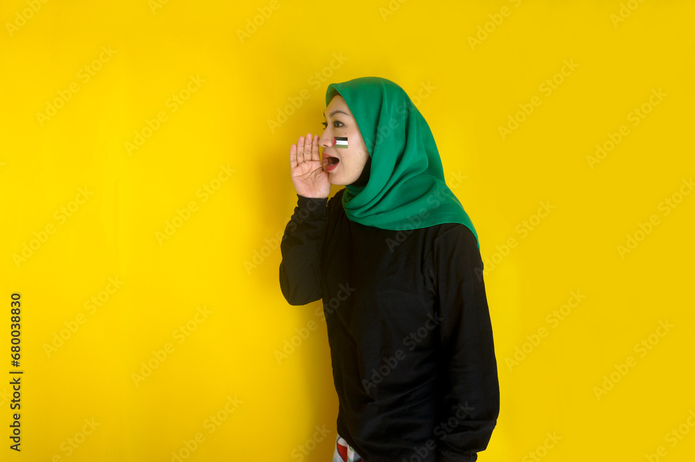 Young Asian muslim woman with Palestine flag on cheek put hand near mouth making announcement gesture Young Asian muslim woman with Palestine flag on cheek put hand near mouth making announcement gesture