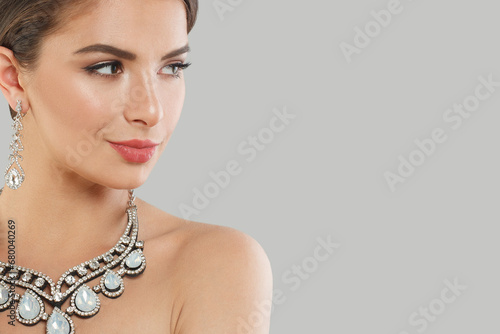 Nice jewelry model woman with dark brown hair and shiny fresh skin wearing diamond earring and necklace posing against grey studio wall background, fashion beauty portrait