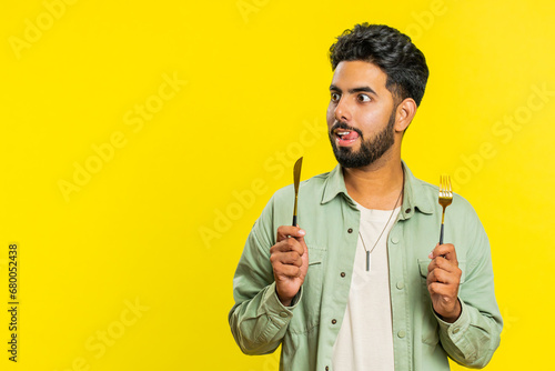 Ready to eat. Hungry Indian man waiting for serving dinner dishes with with restlessness holding cutlery fork knife will appreciate delicious restaurant meal. Excited guy isolated on yellow background photo