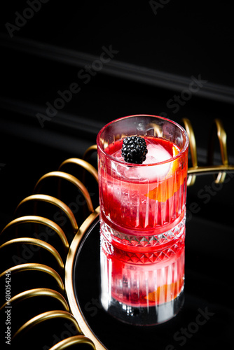 red cocktail in a transparent glass with round ice, whipped cream and black berry on top, side view