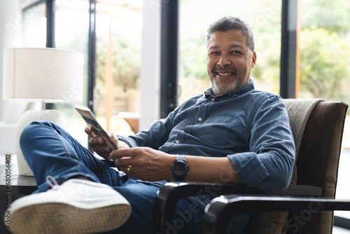 Portrait of happy senior biracial man sitting in armchair and reading book at home photo