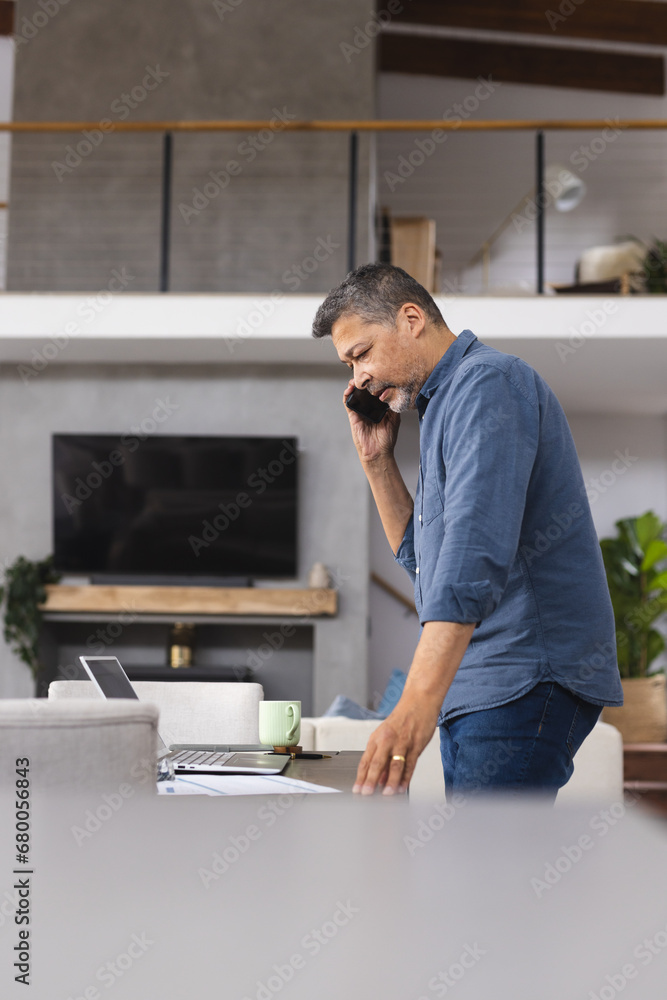 Busy senior biracial man talking on smartphone and working on laptop at home Busy senior biracial man talking on smartphone and working on laptop at home