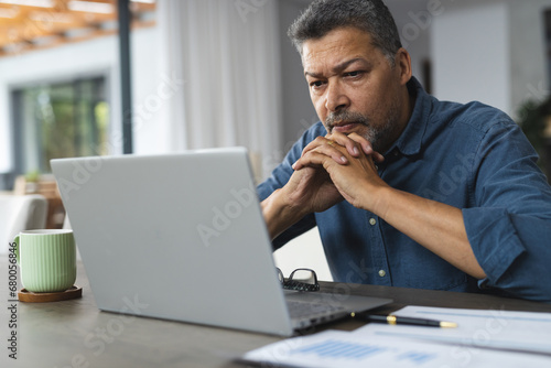 Busy senior biracial man thinking and working on laptop at home