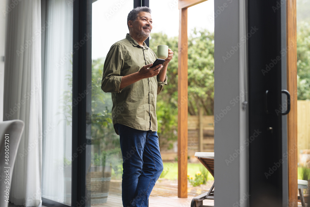 Happy senior biracial man using smartphone at window with cup of coffee at home Happy senior biracial man using smartphone at window with cup of coffee at home