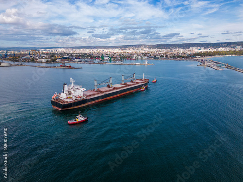 A massive cargo ship bulk carrier leaves the seaport, accompanied by tugboats, aerial view