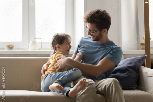 Loving cheerful young father sit on cozy sofa with little daughter at home, play, spend time together. Cute girl tickling laughing handsome father, have fun in living room. Relationship, fatherhood