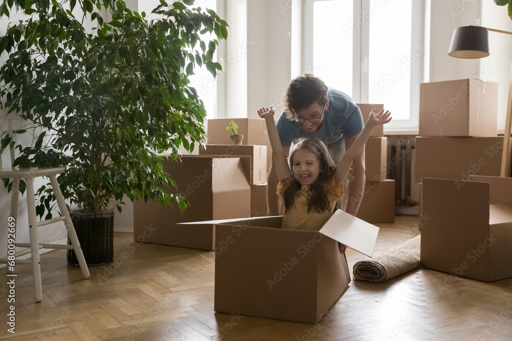 Fototapeta premium Merry little girl sit in big cardboard box play with joyful daddy at new home, dad push forward box with kid inside, have fun together on moving day. Happy family celebrate relocation at own apartment