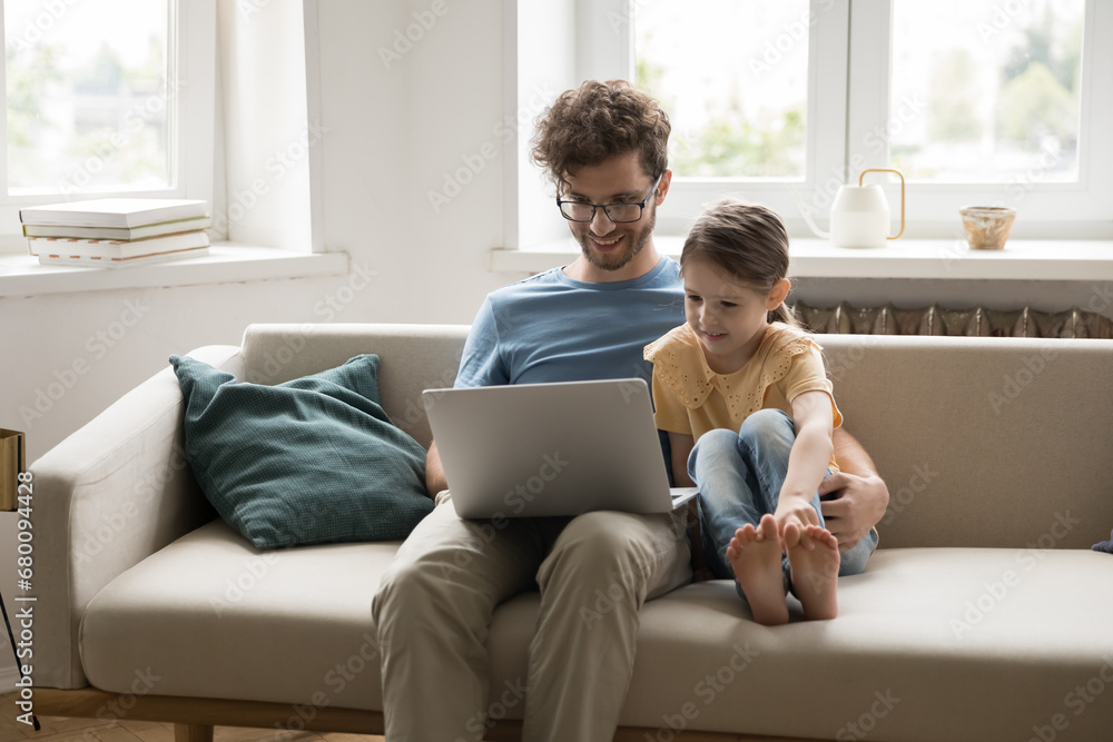 Young father and little daughter relax on cozy sofa spend weekend time at home, look at laptop screen watch funny video, choose cartons. Happy two generation family use modern tech for entertainment