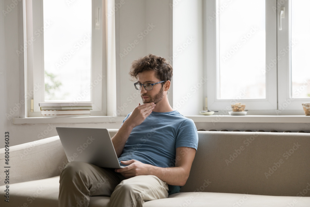 Thoughtful man freelancer sitting with laptop on sofa work on project, thinking about difficult tasks touch chin, solve problem. Serious guy read message, looking for inspiration for new ideas at home Thoughtful man freelancer sitting with laptop on sofa work on project, thinking about difficult tasks touch chin, solve problem. Serious guy read message, looking for inspiration for new ideas at home