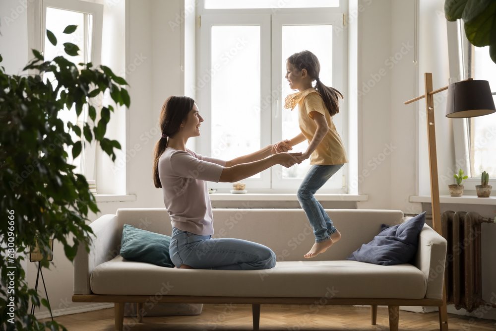 Cheerful mother play with little cute daughter on couch, mom holding hands while joyful girl jumping on soft sofa. Happy family spend time at home, enjoy warm relationship, have fun. Motherhood, love Cheerful mother play with little cute daughter on couch, mom holding hands while joyful girl jumping on soft sofa. Happy family spend time at home, enjoy warm relationship, have fun. Motherhood, love