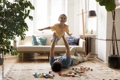 Little girl enjoy playtime with dad at home. In living room young man lying on floor lifts his adorable daughter on outstretched arms, kid pretend flying on air stretched her hands like plane wings photo