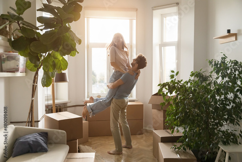 Fotografie Happy husband lifts his young beautiful wife, laughing, swirling, enjoy move-in day to new, first house, big carton boxes nearby in unfurnished sunny living room