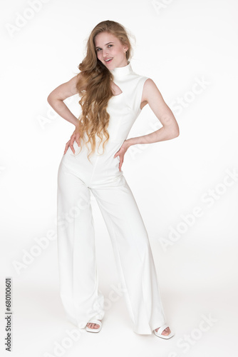 Happy blondie woman with long hair stylishly dressed in white long jumpsuit flowy fabric standing in full length, front view. Smiling Caucasian woman looking at camera, arms akimbo on white background © Alexander Piragis