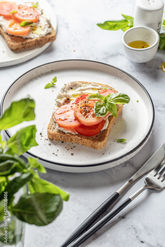 Sandwich or toast with tomatoes, cream cheese, olive oil and basil on a plate on white marble background. Traditional italian mediterranean food