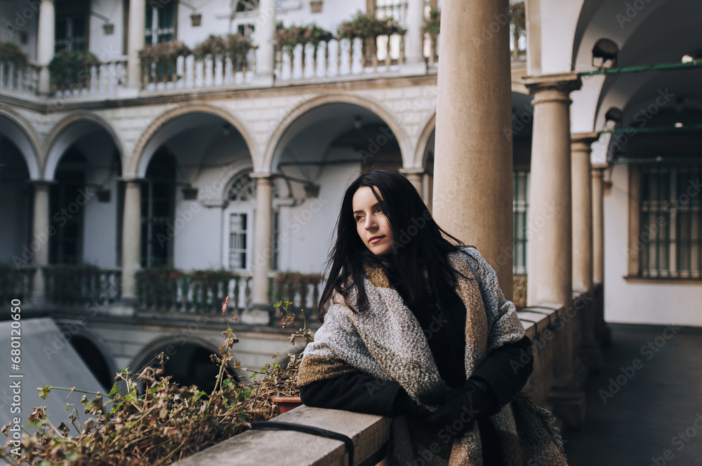 Φωτογραφία A young black-haired girl in a dark autumn coat and a wide scarf-wrap stands on the balcony with a colonnade and looks down thoughtfully