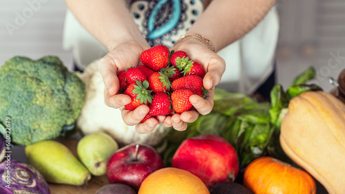 Female hands with fresh strawberry above vegetables and fruits on a white table in the kitchen, close-up, selective focus.