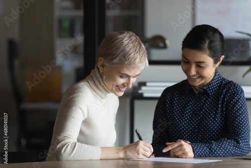 Happy diverse businesswomen signing contract, loan, insurance agreement. Lawyer helping customer to fill document form, writing, pointing at paper. Paperwork, legal expertise, consultation concept photo