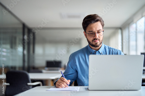 Young busy latin business man employee worker looking at laptop computer watching online webinar or having virtual hybrid meeting writing notes, doing market research working in office. Copy space.