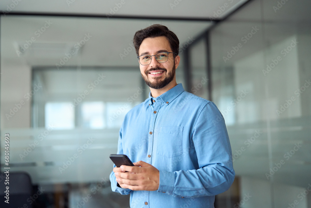Naklejka premium Happy young latin business man holding smartphone standing in office. Smiling hispanic businessman entrepreneur or manager using financial banking apps on cell phone technology device at work.