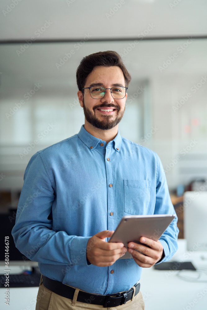 Smiling young latin business man manager using tablet computer, happy hispanic businessman employee, company worker looking at camera holding tab working standing in office. Vertical portrait.