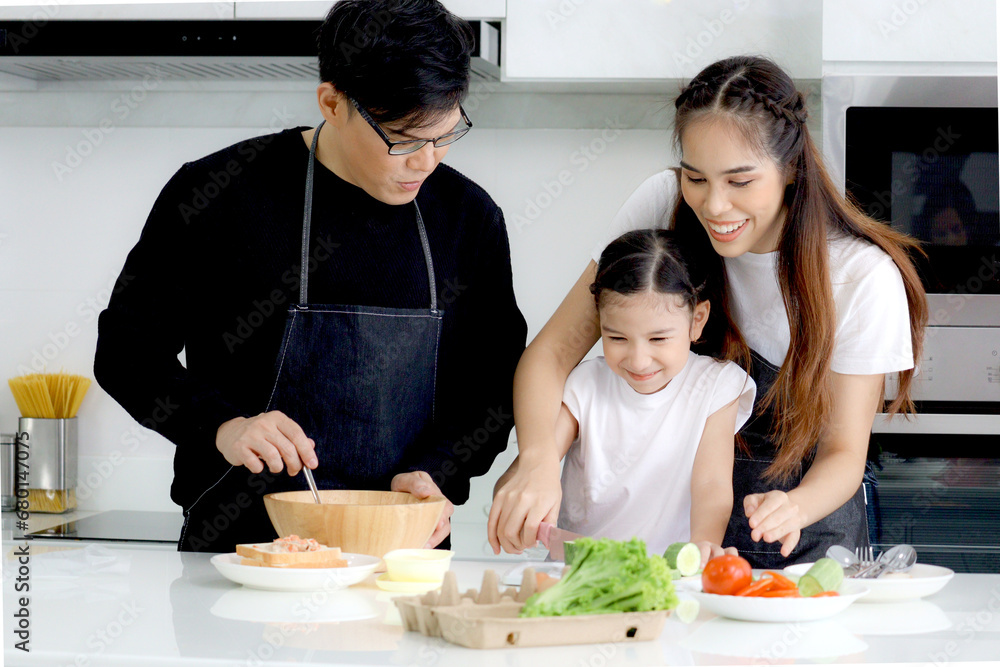 Happy family cooking at kitchen, father mother and cute daughter girl having fun during making meal at home. Parents and kid child enjoy cooking food, spending time together. Family love and bonding.