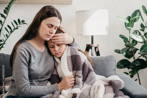 Caring mum comforting sad ill kid, expressing love and support, child psychologist concept. Young caucasian mother hugging sick upset little daughter covered blanket, sitting together on sofa at home