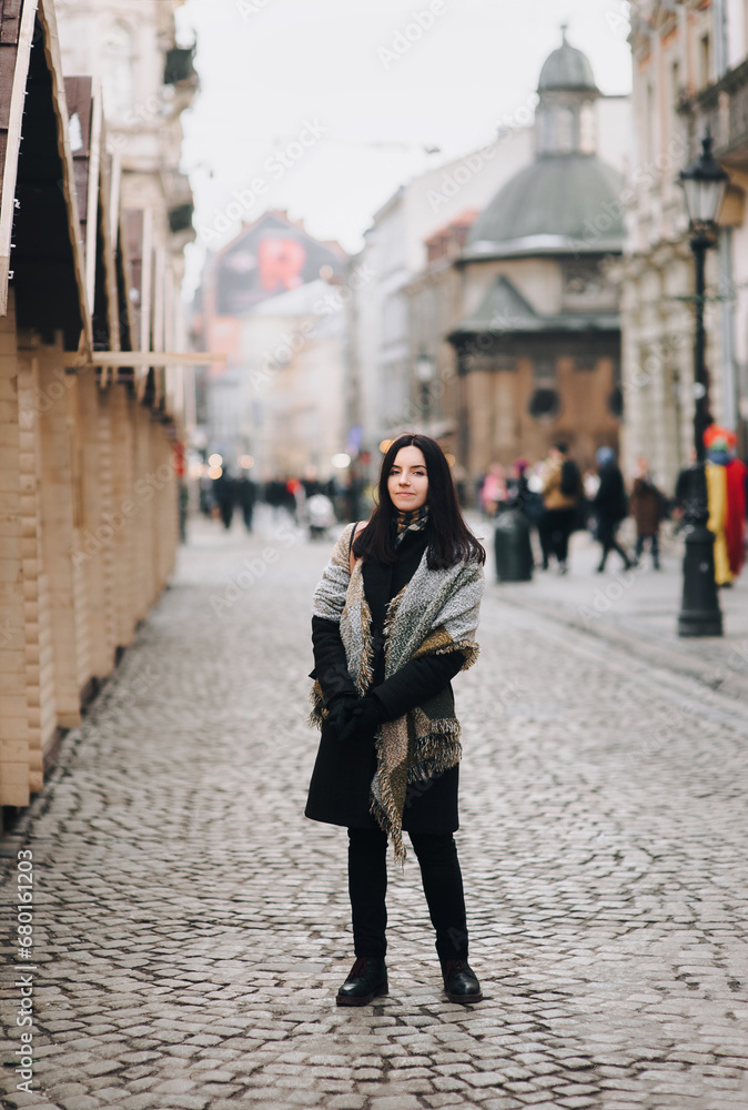 A young girl in a dark coat and a wide scarf stands against the backdrop of a street with passers-by. Lviv, Ukraine.
