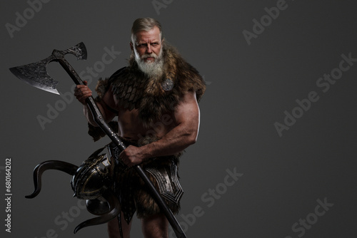 Aged bearded Viking warrior dressed in fur and light armor, holding a two-handed axe, against a grey background