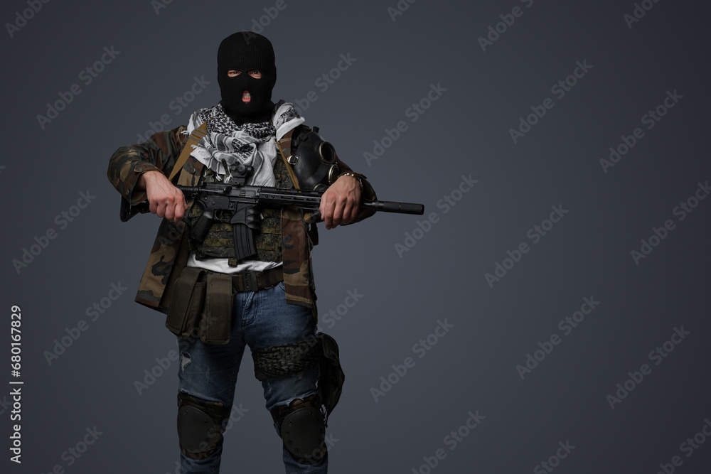 Portrait of a Middle Eastern radical soldier wearing a black balaclava and camouflaged field uniform, armed with an automatic rifle, against a cold gray background