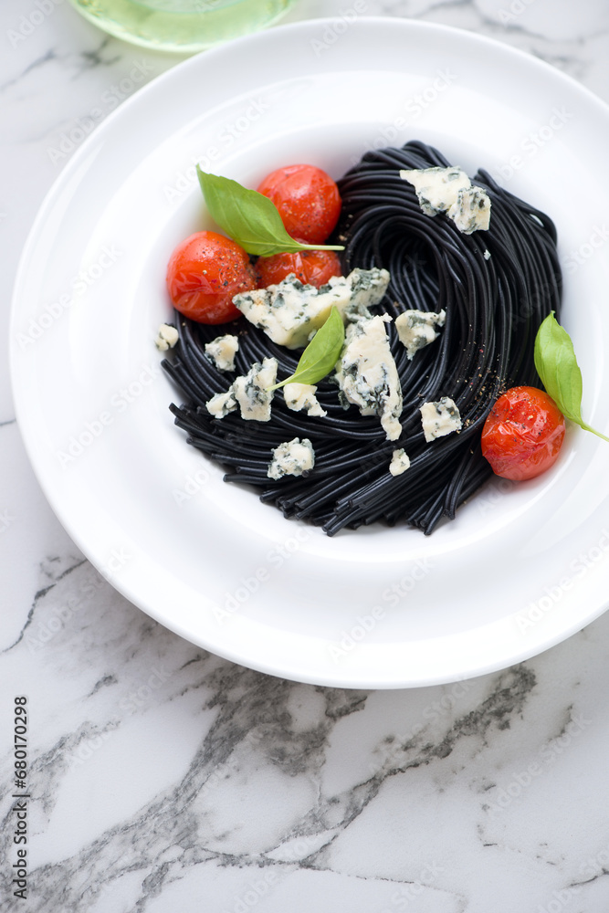 Plate with black spaghetti, blue cheese, roasted tomatoes and basil, vertical shot on a white marble background, elevated view Plate with black spaghetti, blue cheese, roasted tomatoes and basil, vertical shot on a white marble background, elevated view