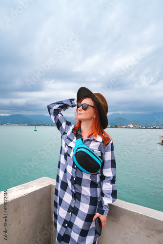 Young woman having a rest at the seacoast. Black Sea landscape. She is touching her hat. Front view. sunglasses and small bag. Travel and vacation. Vertical photo. Batumi Georgia