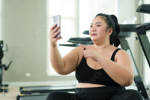 Chubby woman sitting and resting after exercising in fitness center. Video call with friends via smartphone To invite you to exercise The joy of a cute fat Asian woman trying to lose weight